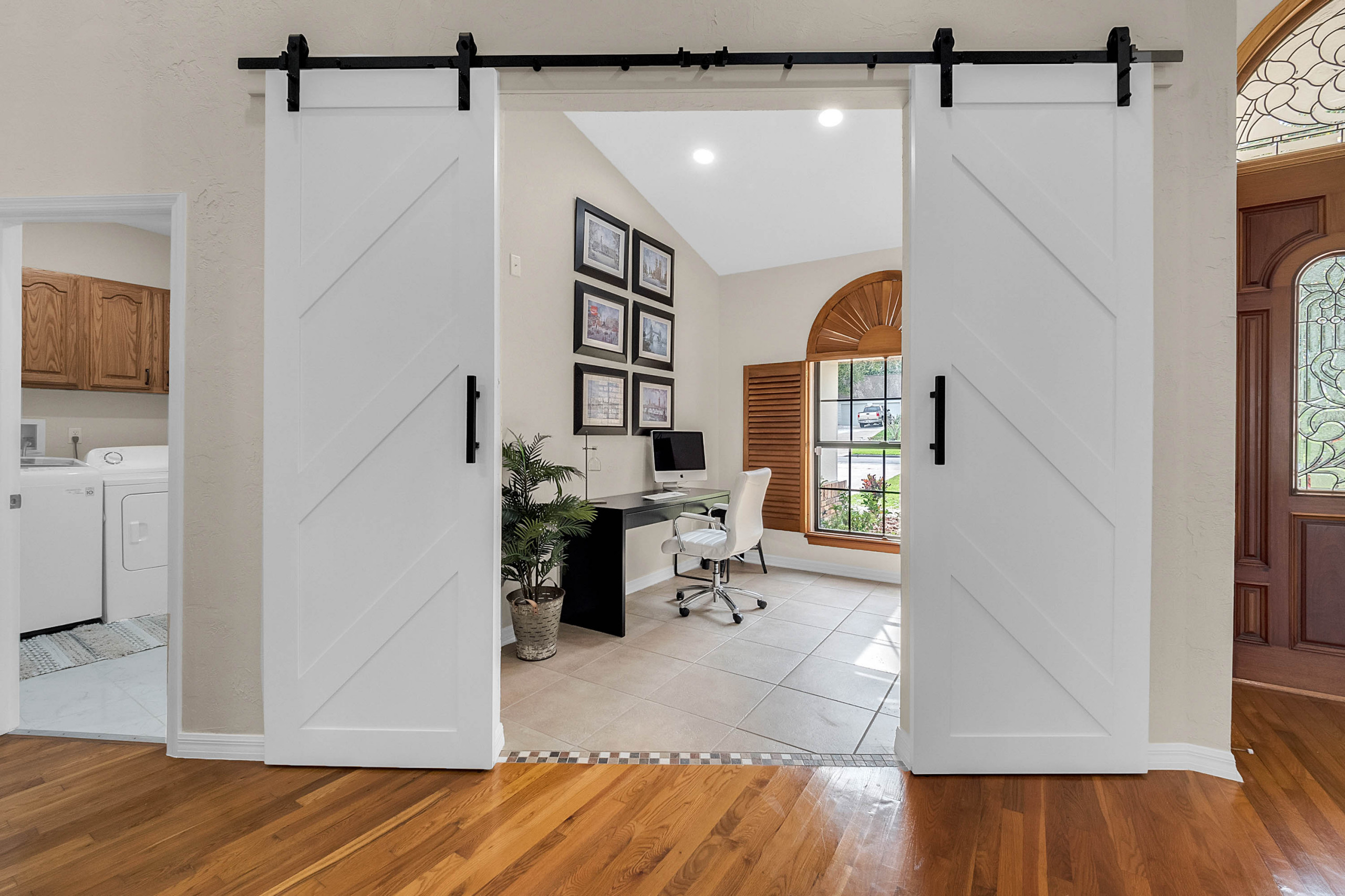 White barn doors open to a neutral home office with a black desk, white chair, and framed art.