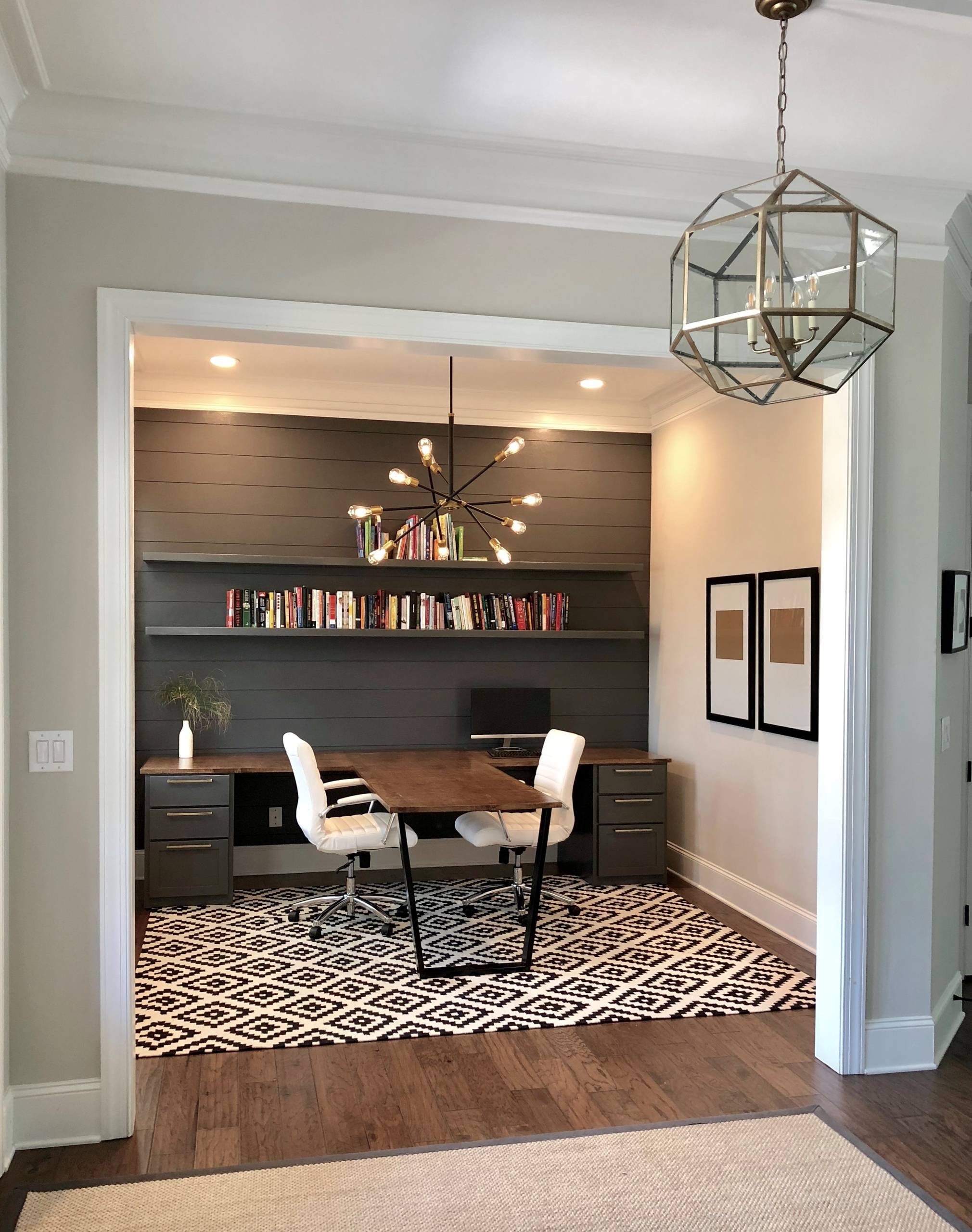 Modern home office with dark shiplap wall, light wood desk, white chairs, and a bold black and white geometric rug.