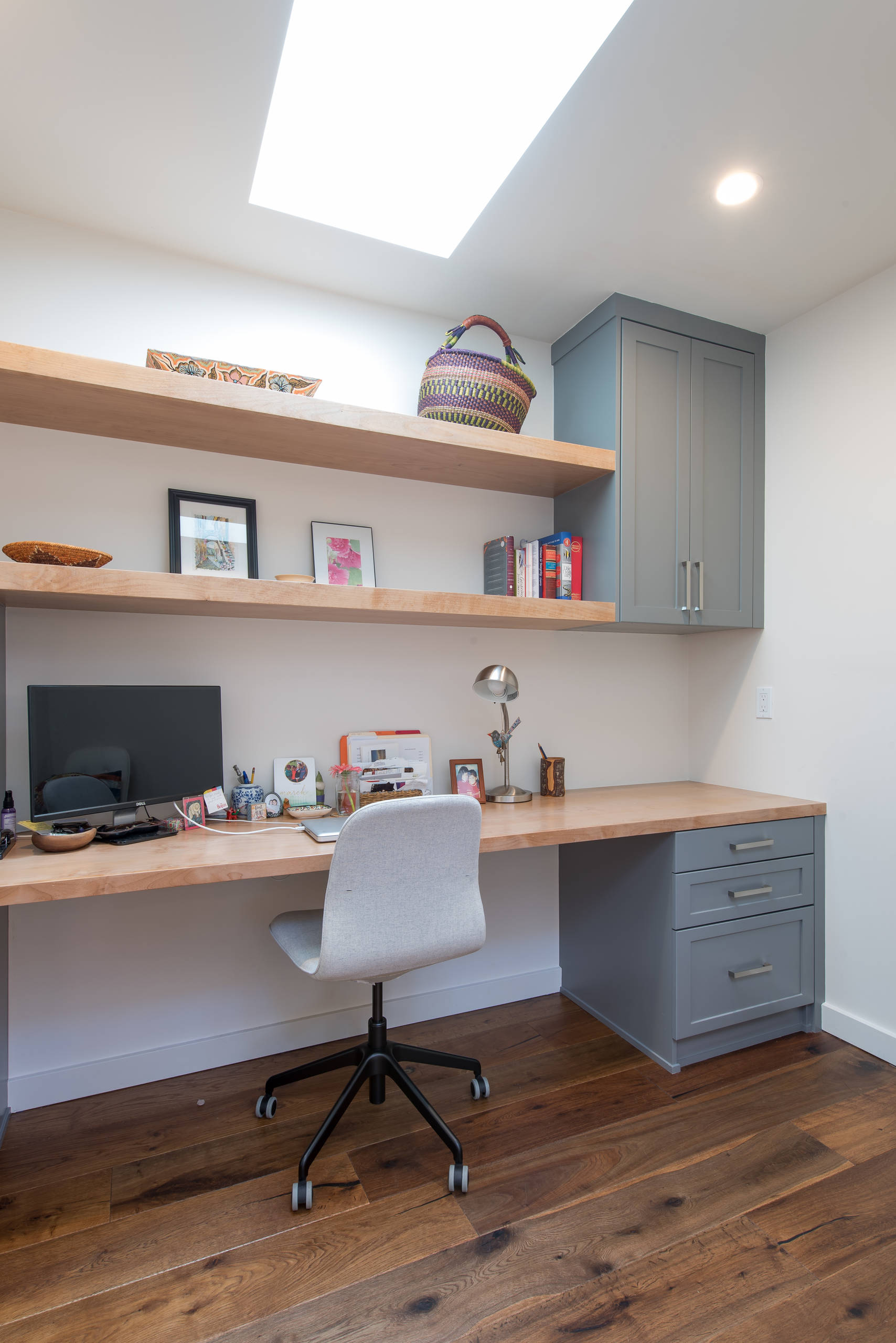 Bright modern home office with light wood desk, grey cabinets, floating shelves, and a large skylight.