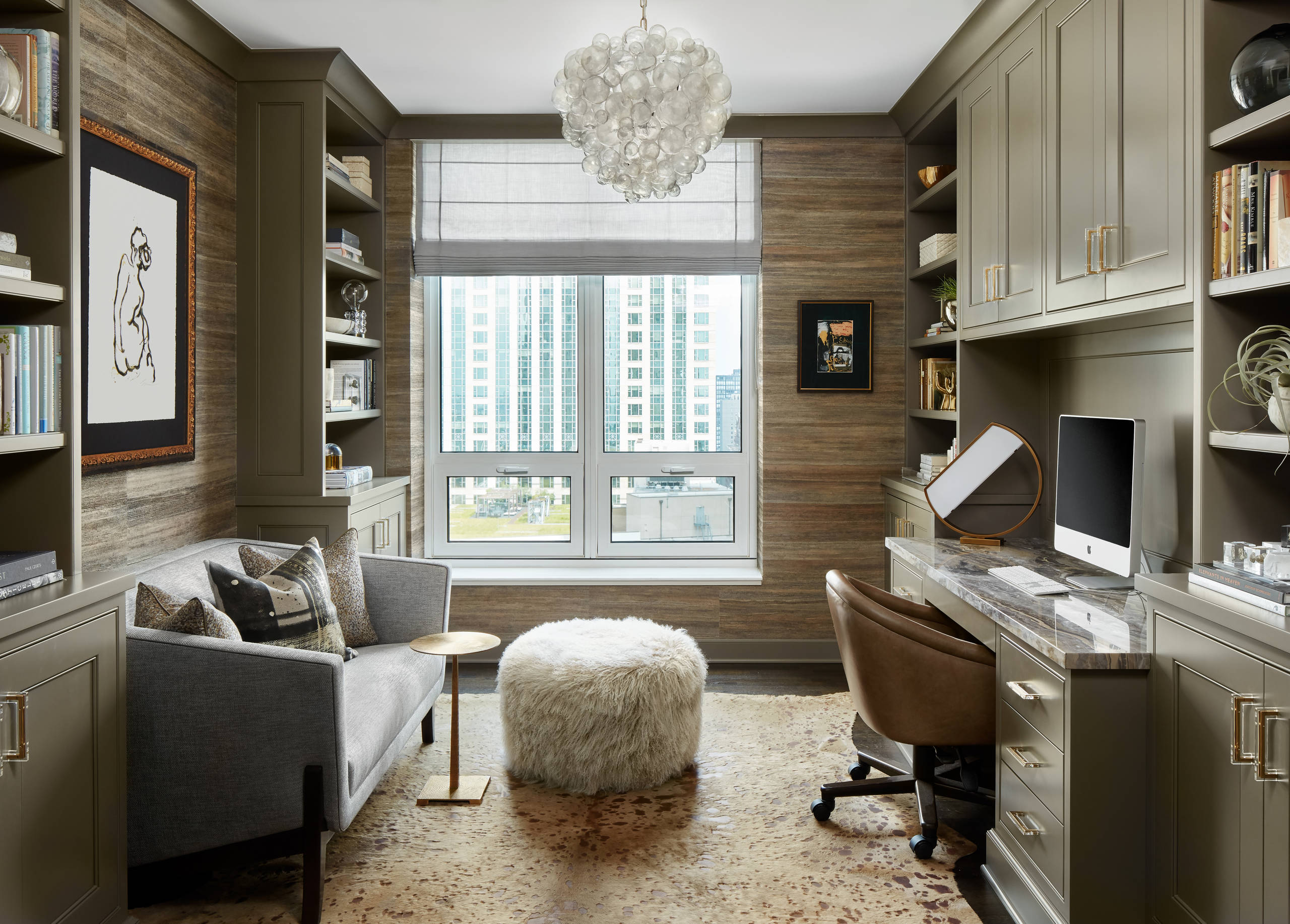 Modern study with olive-gray built-ins, textured wood walls, cowhide rug, gray sofa, marble desk, and city view.