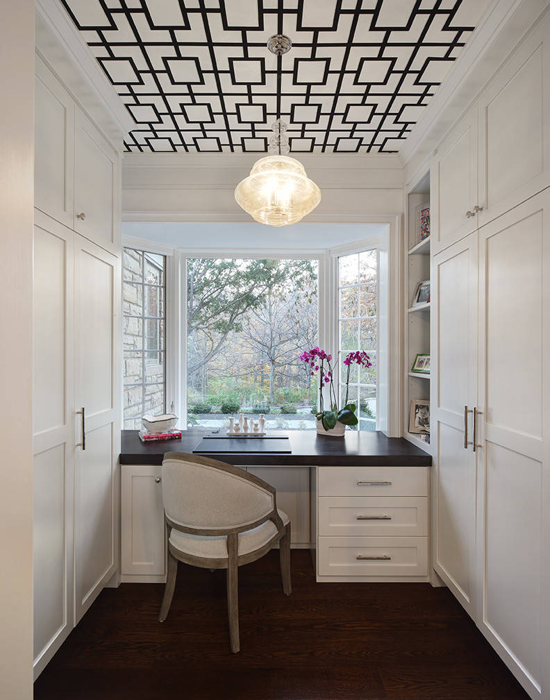 Home office with black geometric ceiling, white built-in cabinetry, dark desk, and bay window overlooking trees.