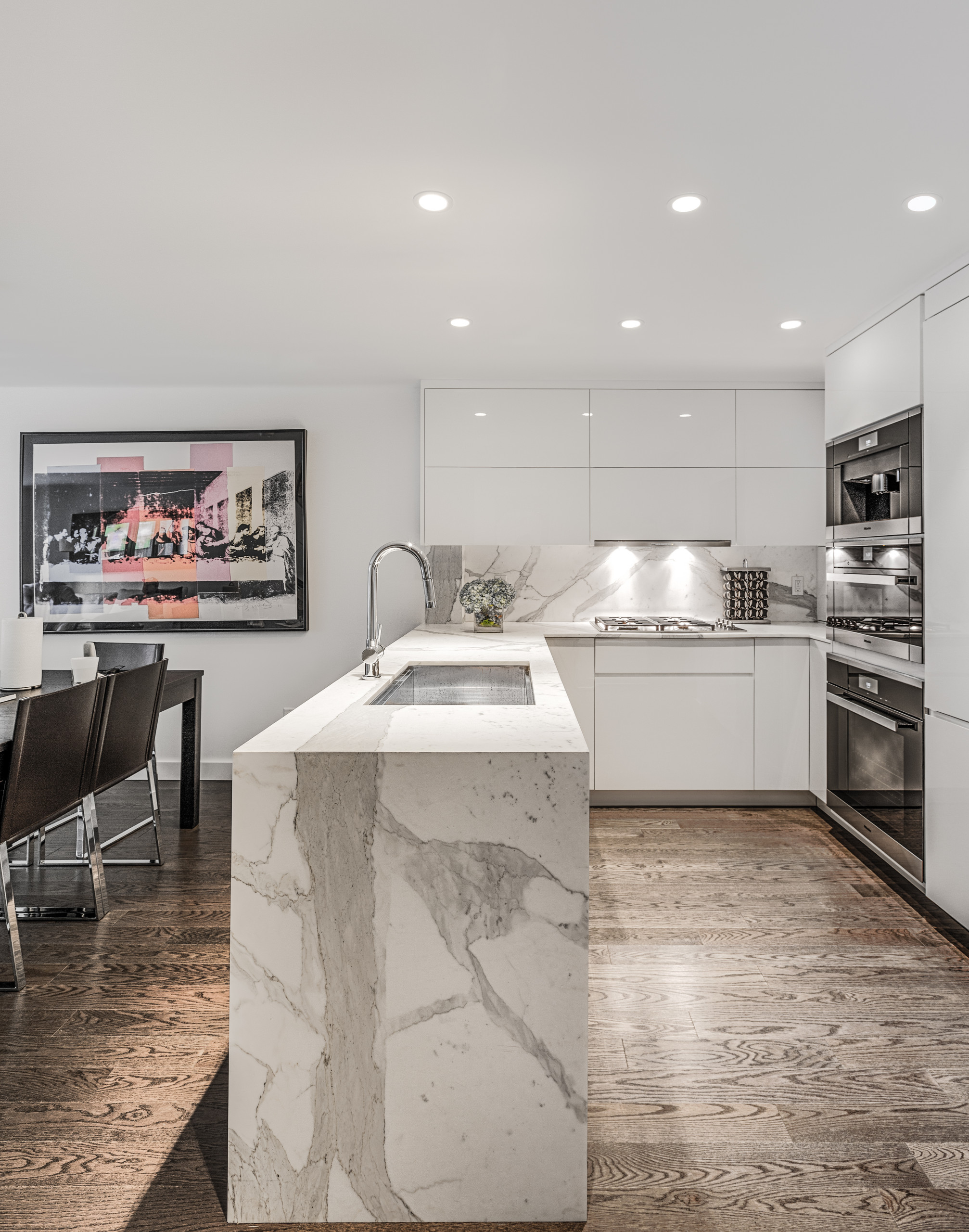 Sleek white modern kitchen with a striking marble waterfall island, dark wood floor, and integrated appliances.