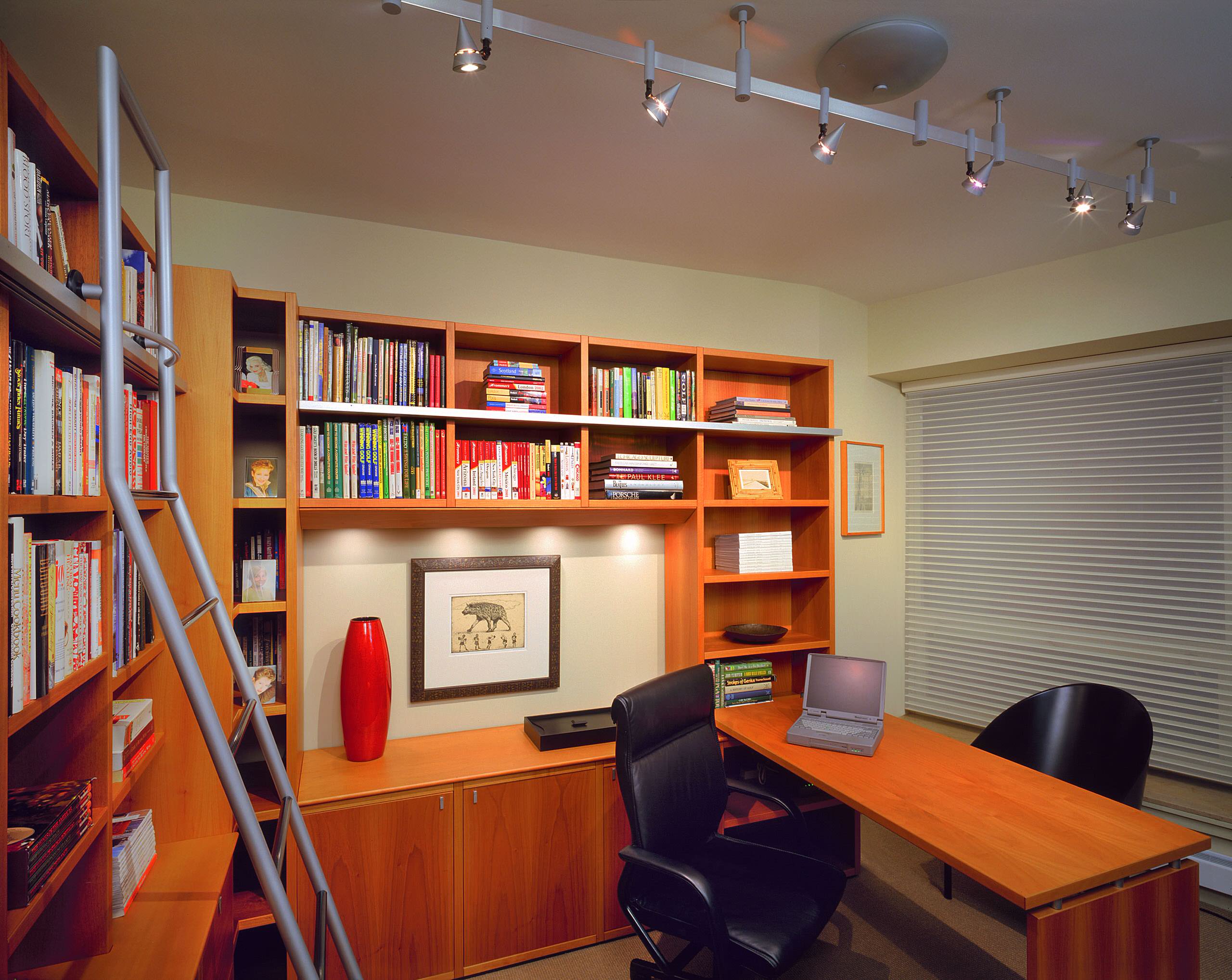 Warm-toned wood study with built-in bookshelves, track lighting, a ladder, and a sleek desk.