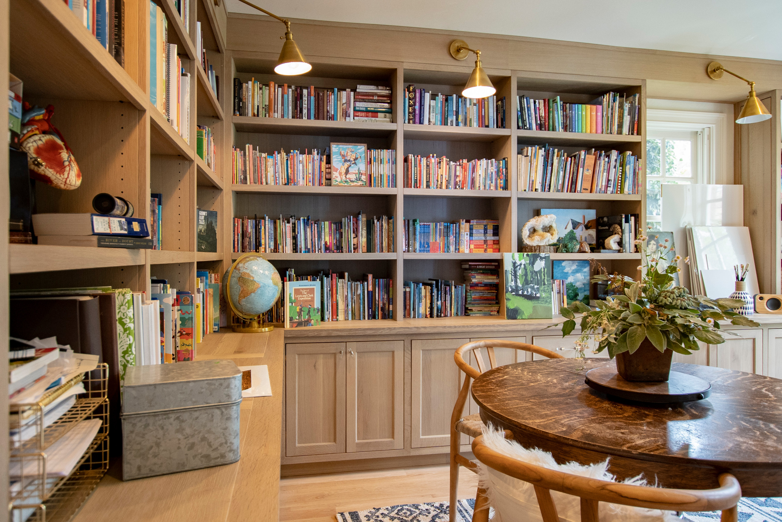 Light-toned wood library office with extensive bookshelves, brass sconces, and a round wooden table.