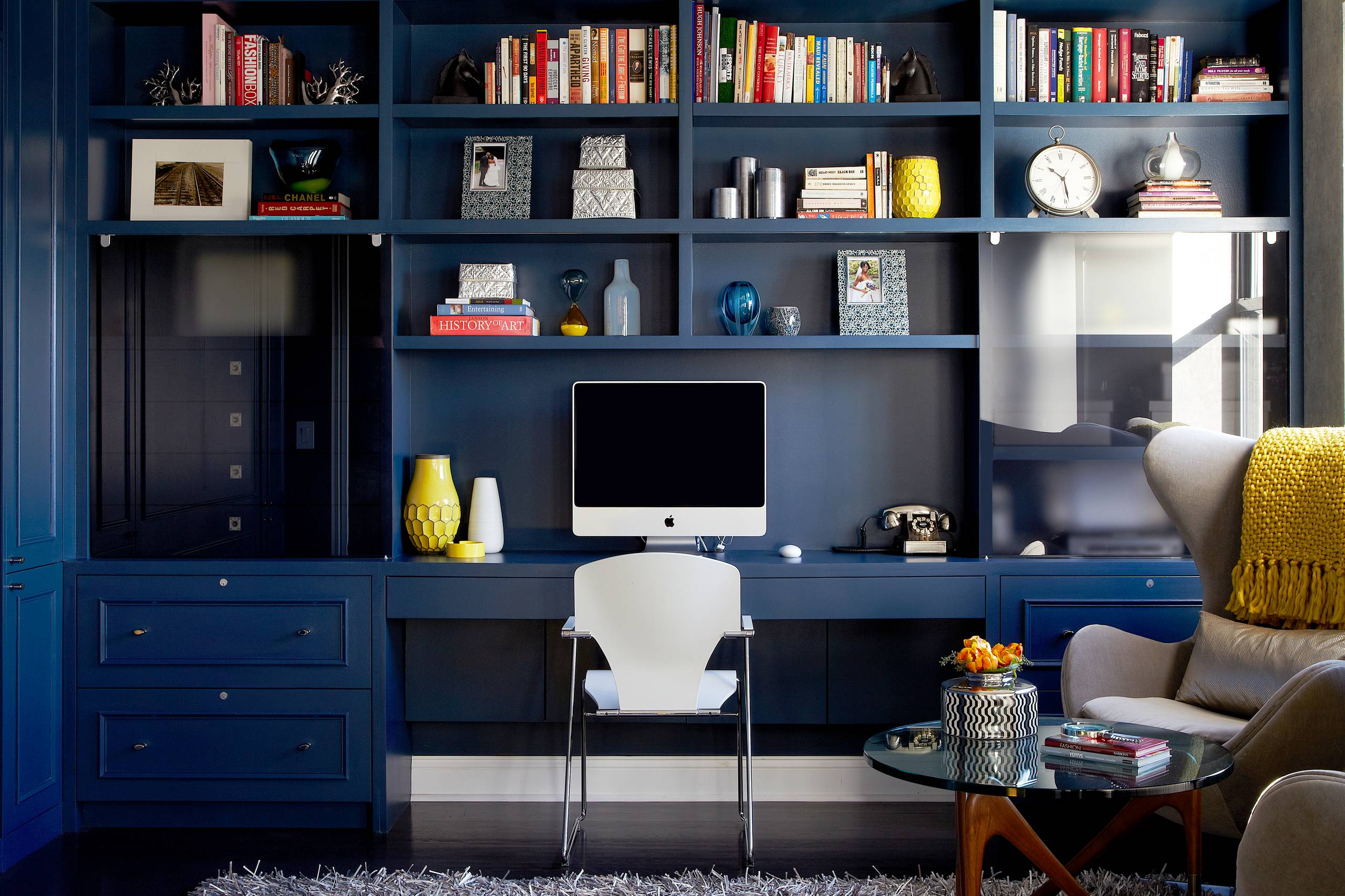 Navy blue custom built-in shelving with integrated desk, white modern chair, and gray armchair with yellow throw.