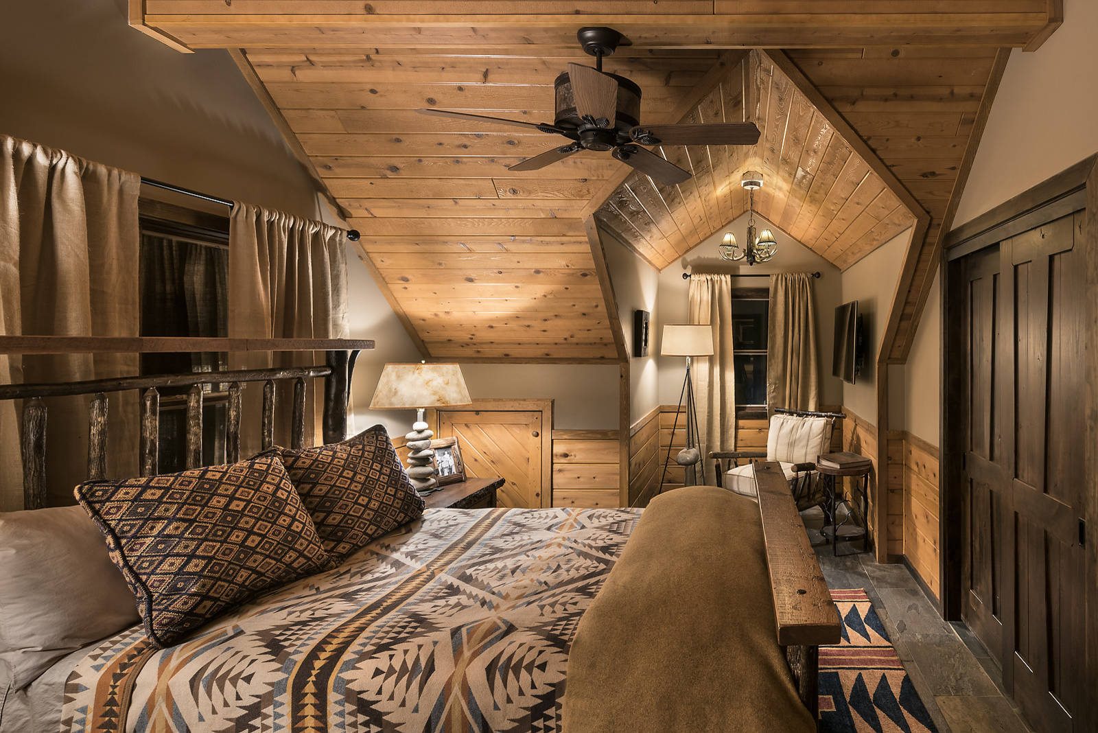 Cozy rustic bedroom with angled wood-paneled ceiling, log bed frame, patterned bedding, and stone floor.