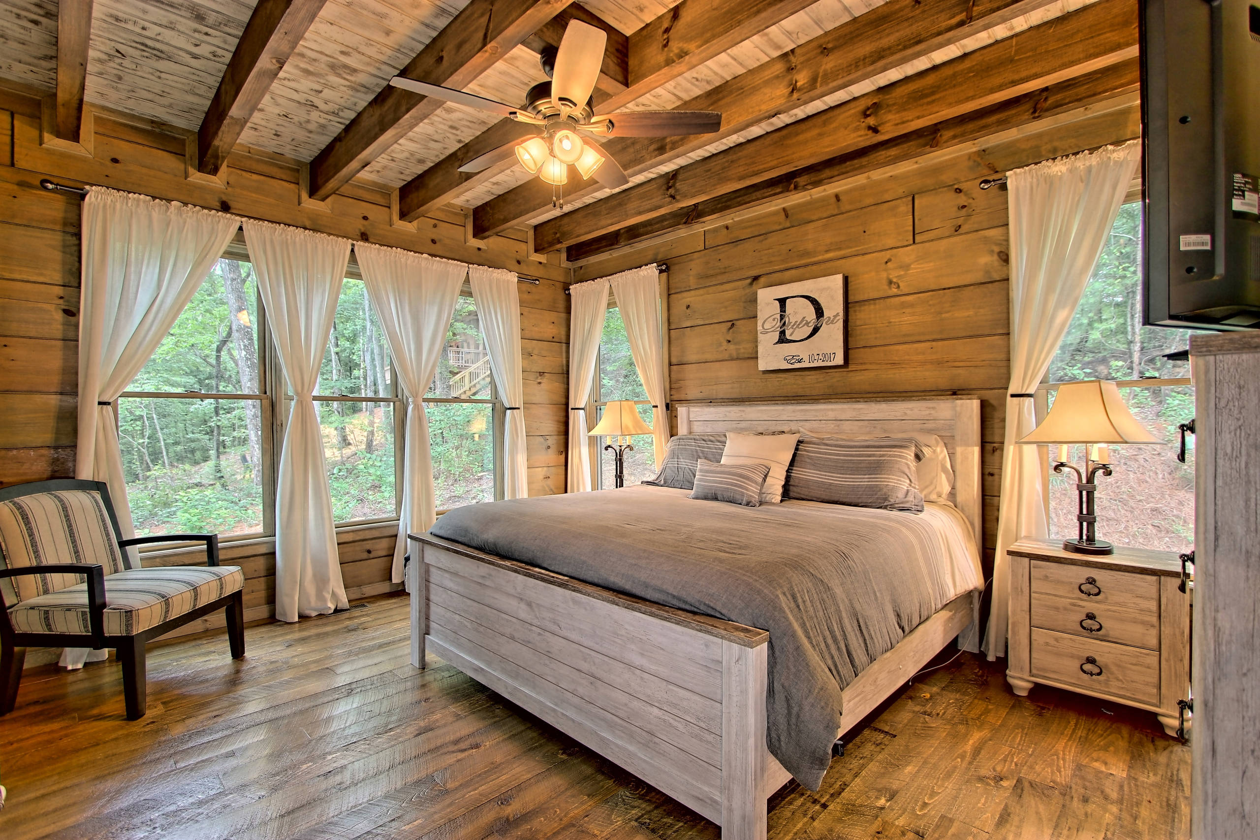 Cozy bedroom with wooden walls, ceiling beams, white bed, sheer curtains, and forest views.