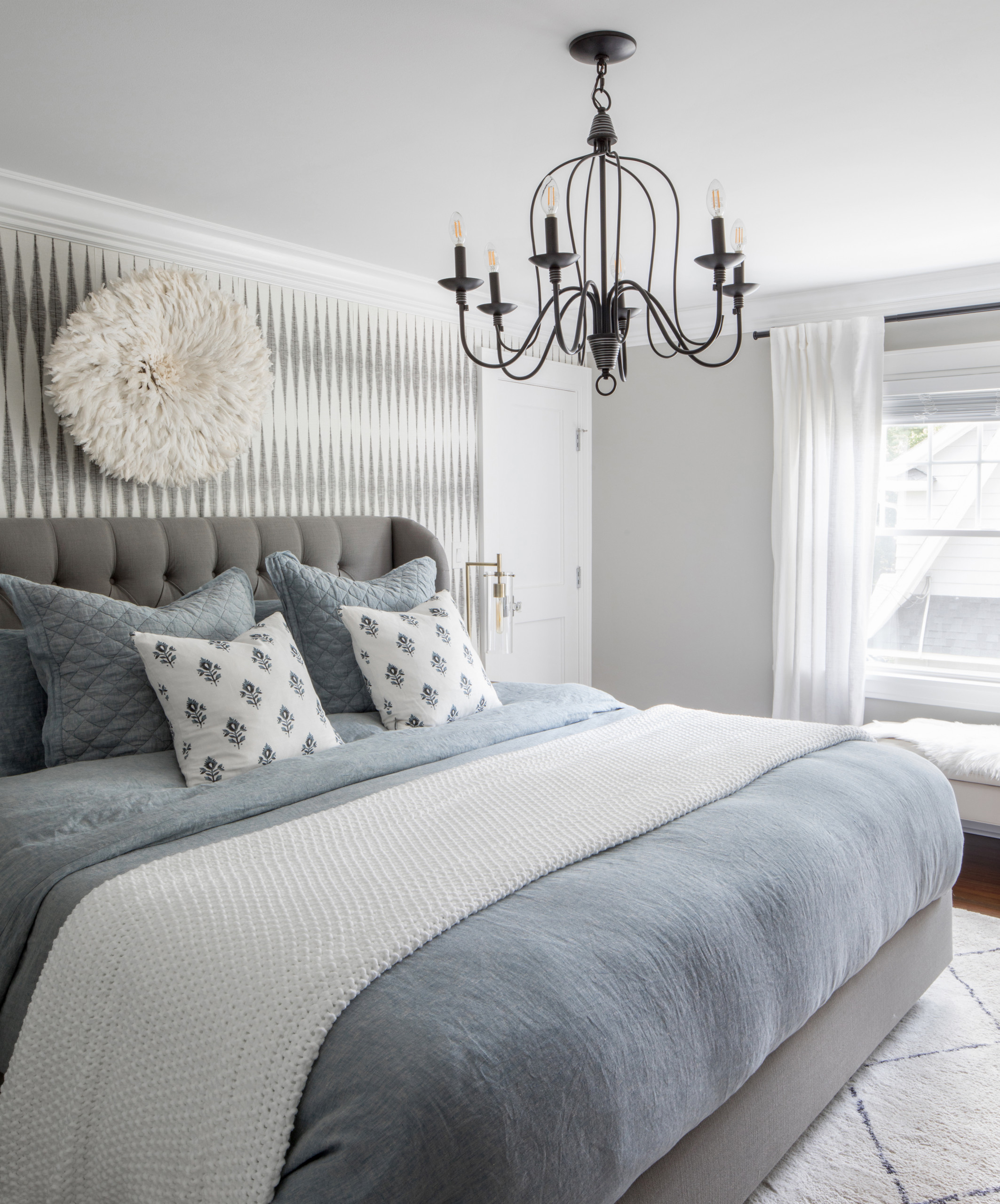 Bedroom with gray tufted headboard, blue bedding, black chandelier, geometric wallpaper, juju hat.