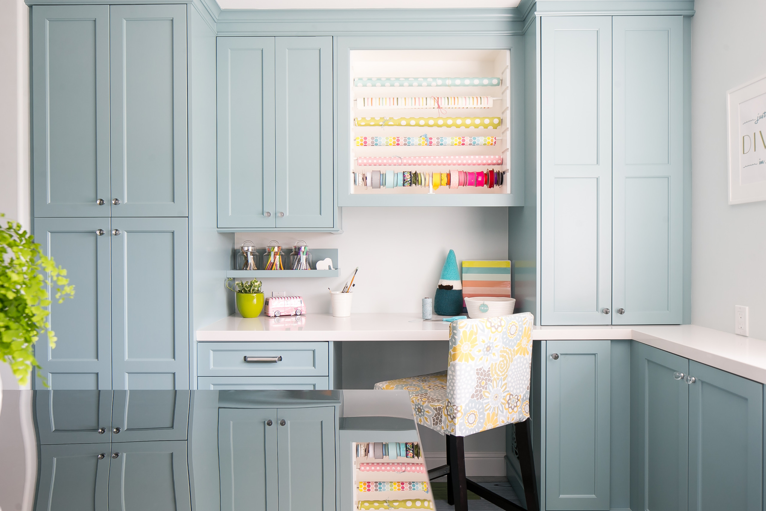 Sky blue craft room with white desk, extensive cabinetry, open storage for wrapping paper, and a floral upholstered chair.