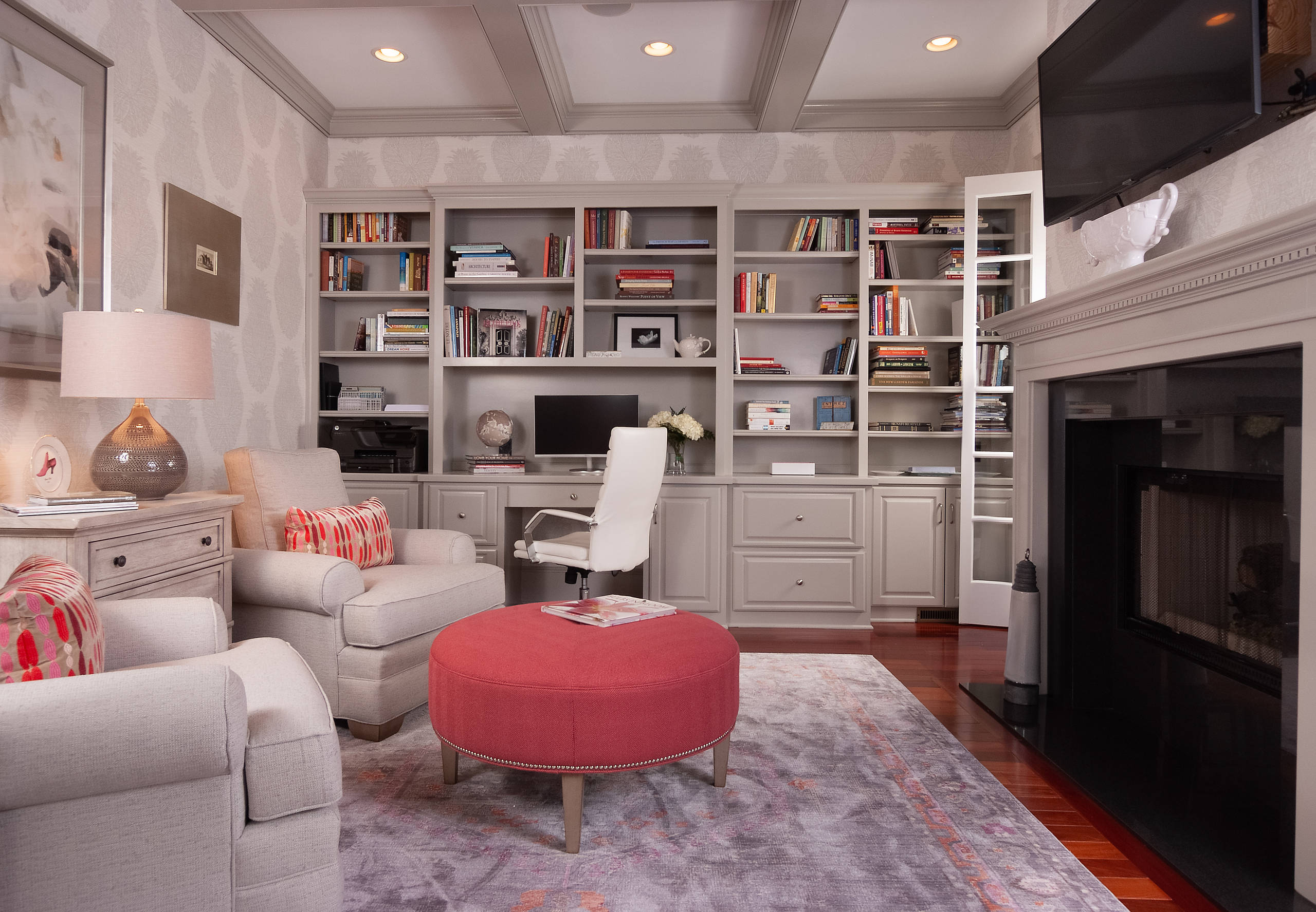 Taupe built-in shelving with integrated desk, white office chair, two beige armchairs, and a red ottoman.