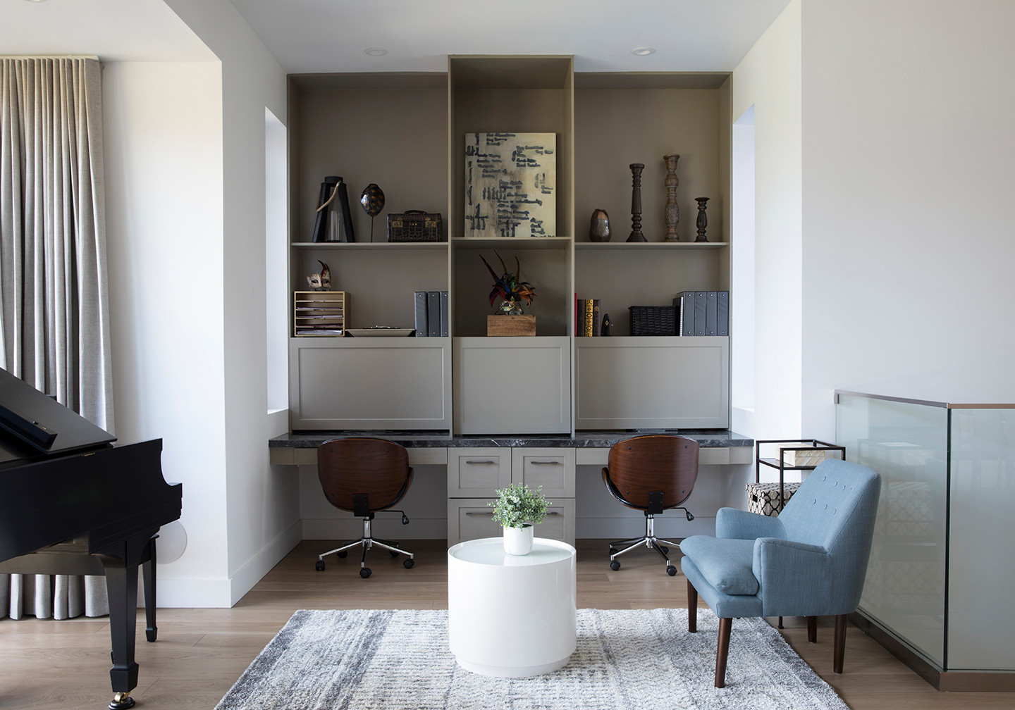 Taupe built-in shelving with two integrated desks, wood chairs, light blue armchair, and baby grand piano.