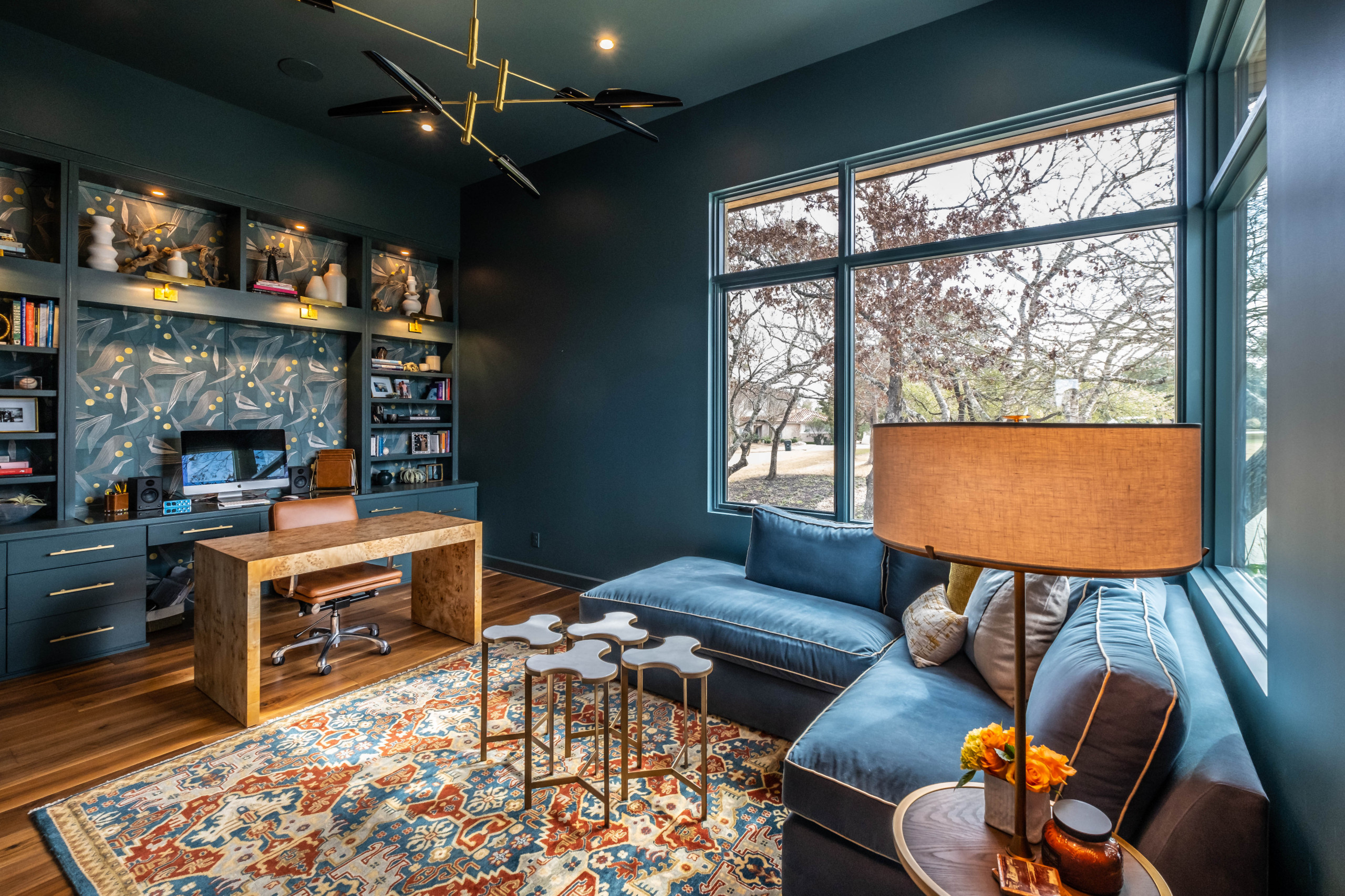 Teal home office with velvet sectional, burl wood desk, and vibrant global patterned rug.