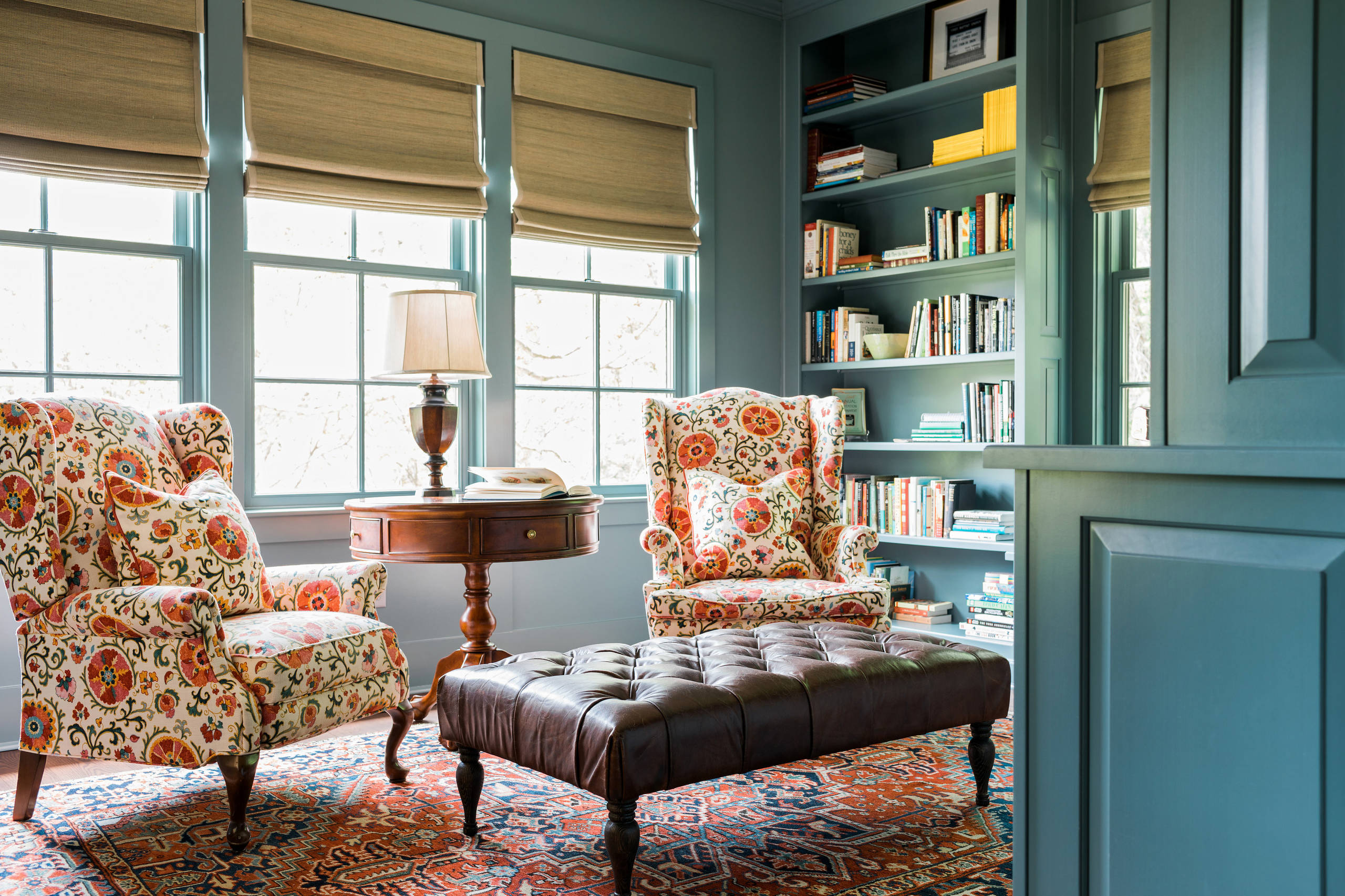 Cozy teal library with two patterned armchairs, round wooden table, leather ottoman, and built-in bookshelves.