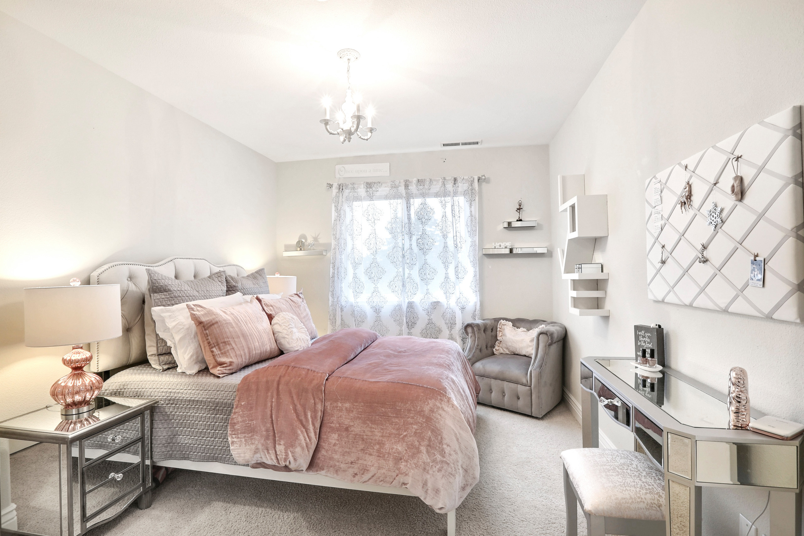 Feminine bedroom with tufted gray bed, pink velvet duvet, mirrored nightstands, vanity, and gray armchair.