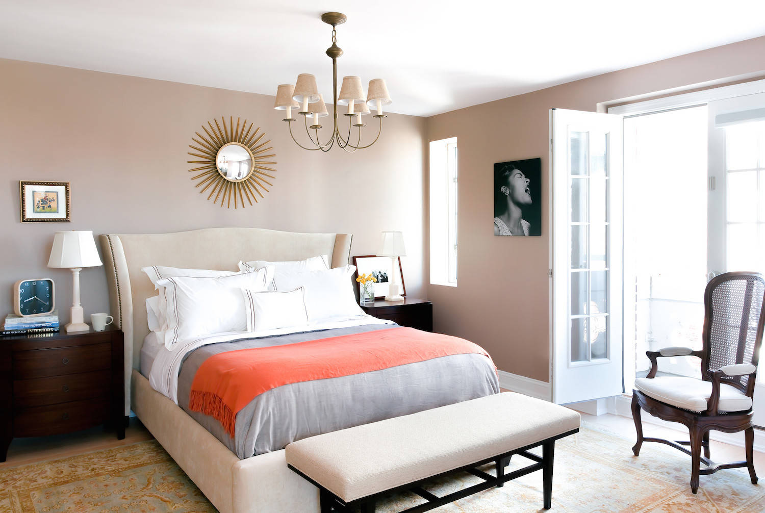 Traditional bedroom with neutral upholstered bed, dark wood nightstands, coral throw blanket, and sunburst mirror.