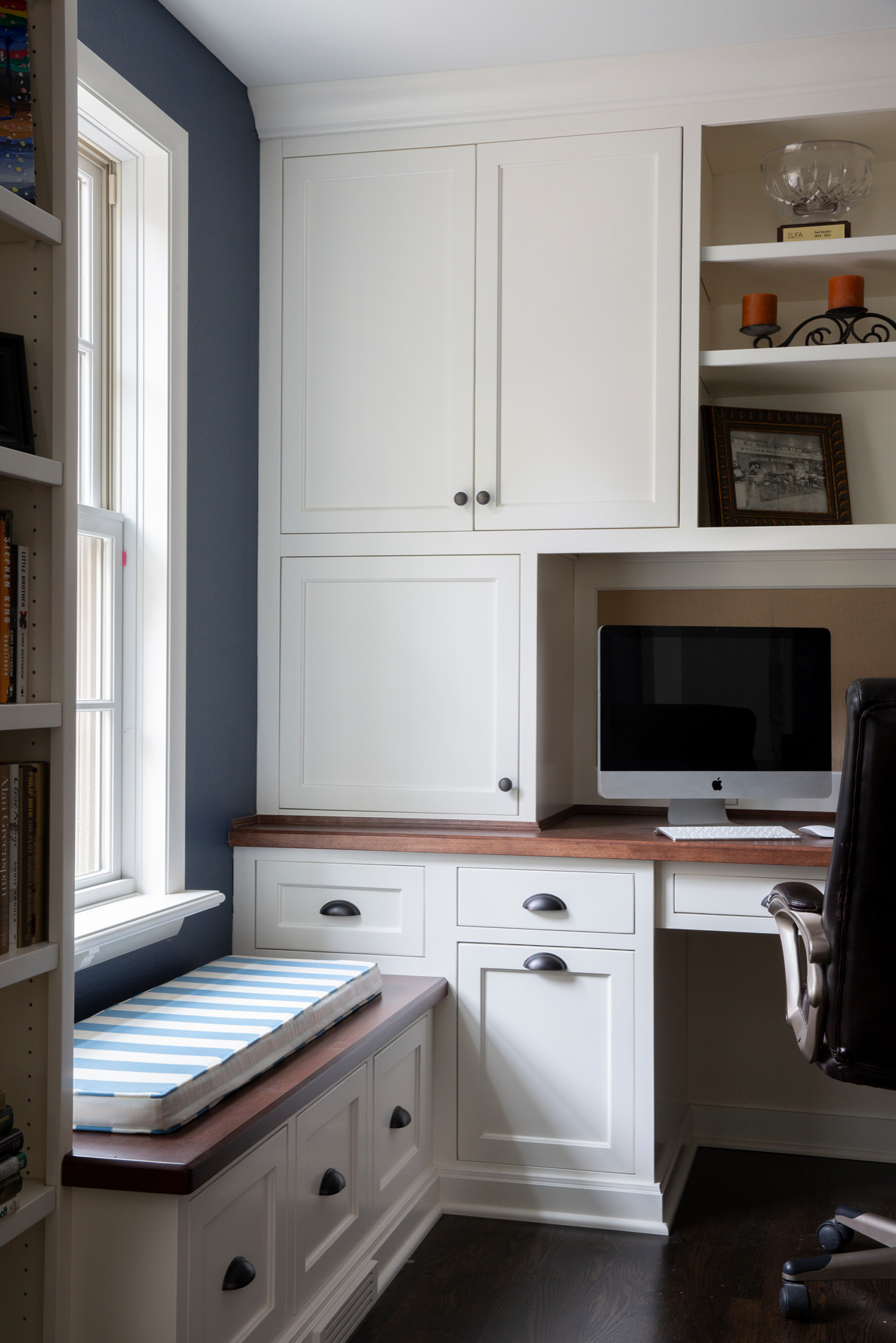 A cozy home office with white built-in cabinetry, dark wood desk, navy accent wall, and window seat.