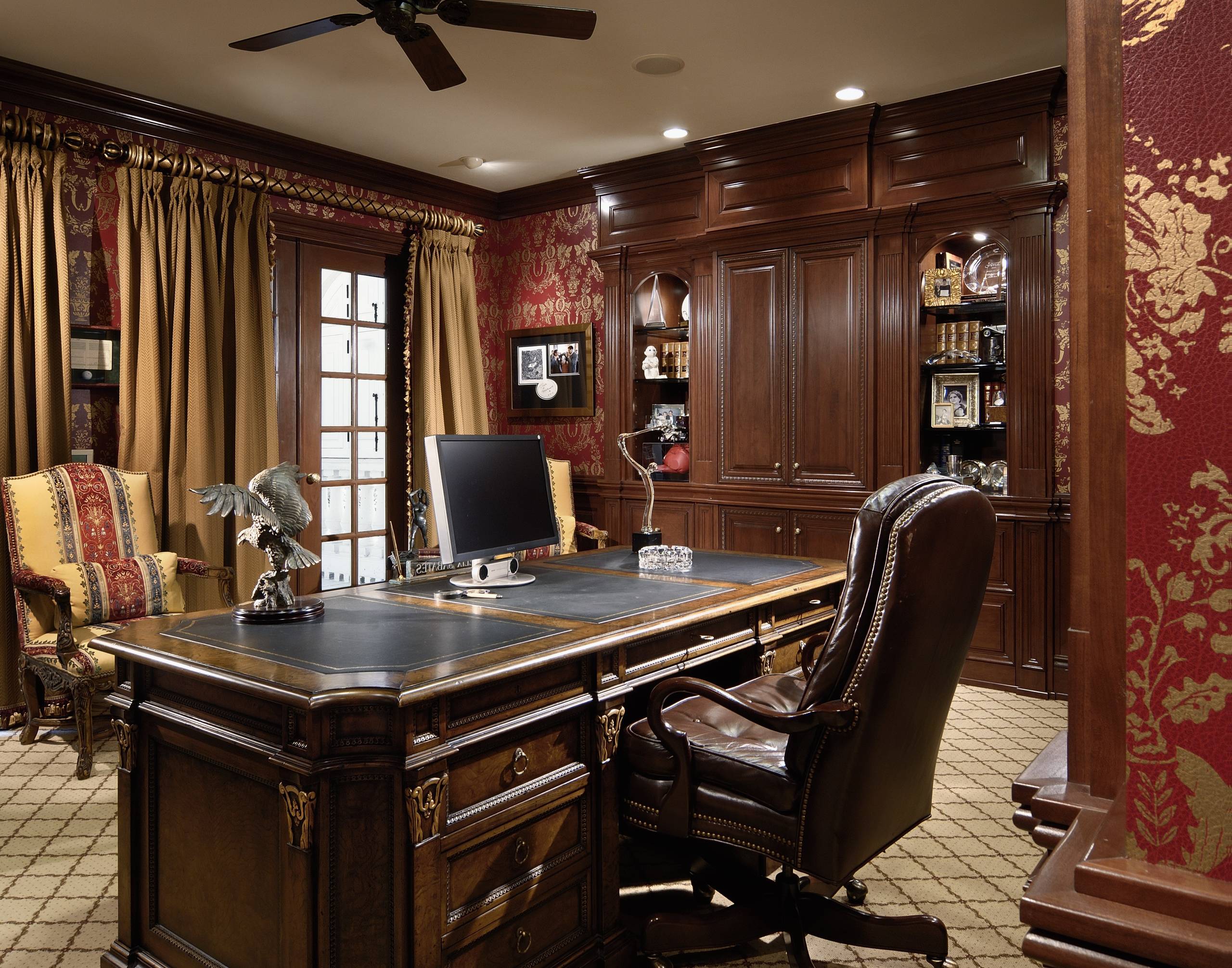 Luxurious traditional home office with dark cherry wood cabinets, red and gold damask wallpaper, leather desk, and patterned rug.