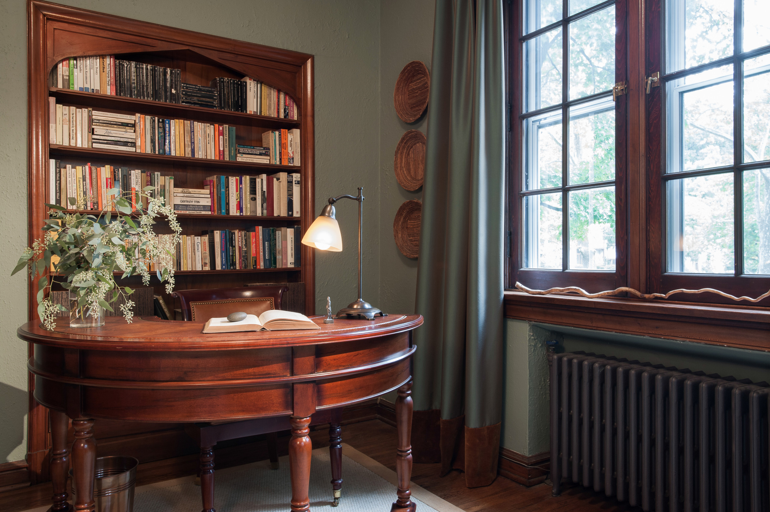A traditional home office with dark wood desk, built-in bookshelf, sage green walls, and large gridded window.