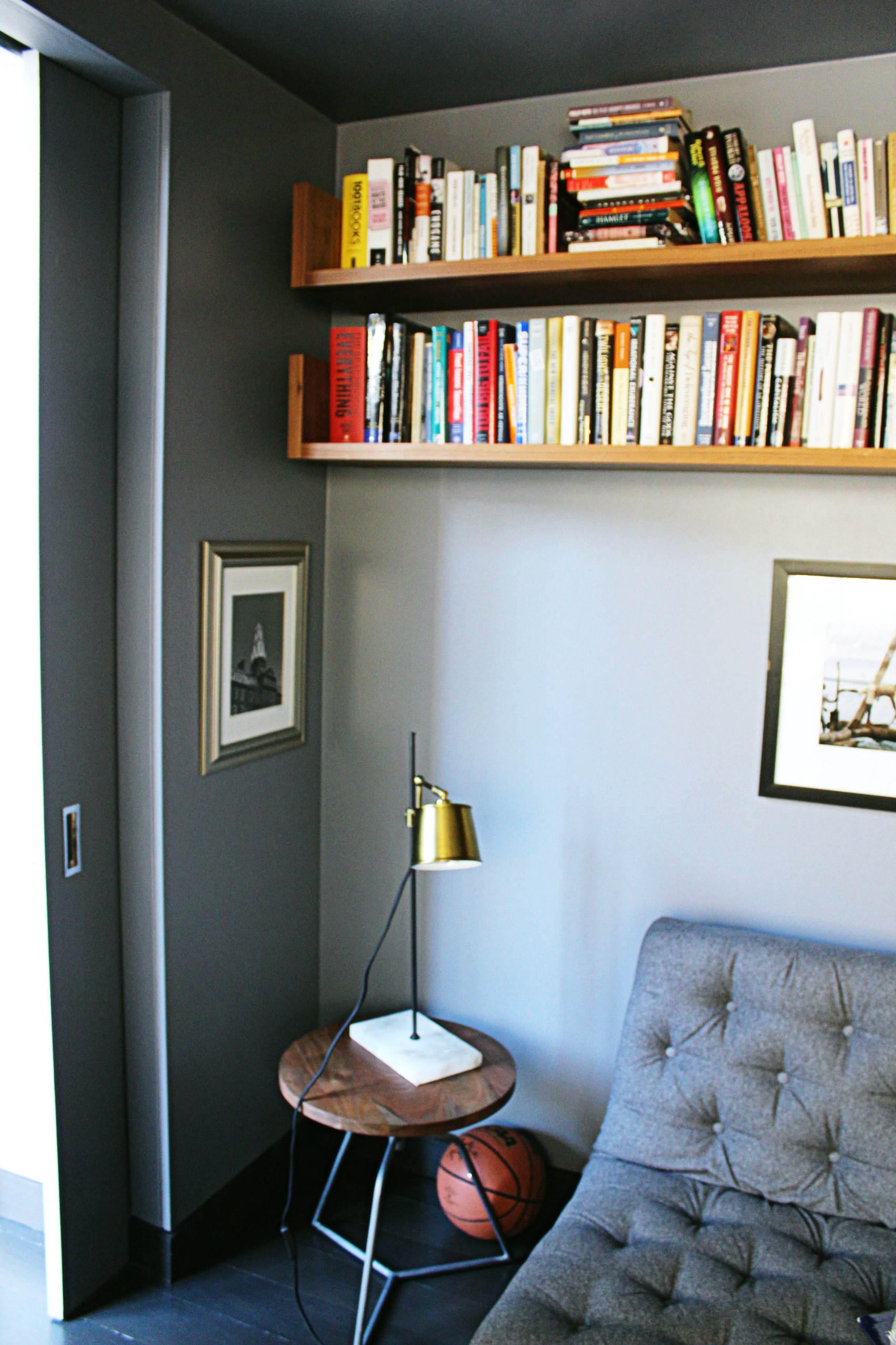 Cozy reading nook with light grey walls, floating wood bookshelves, tufted grey lounge chair, and brass task lamp.