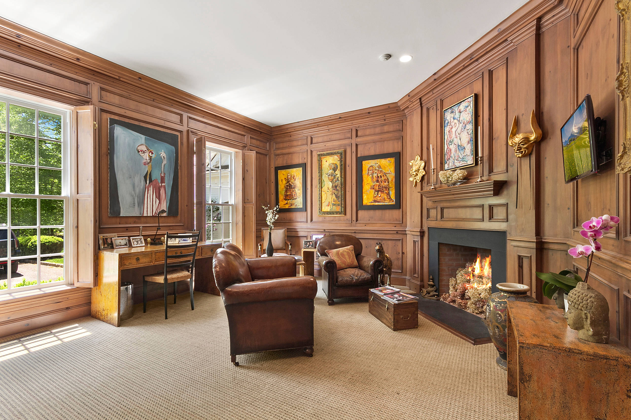 Rich wood-paneled study with leather armchairs, a lit brick fireplace, and vibrant framed art, showcasing a blend of comfort and curated design.
